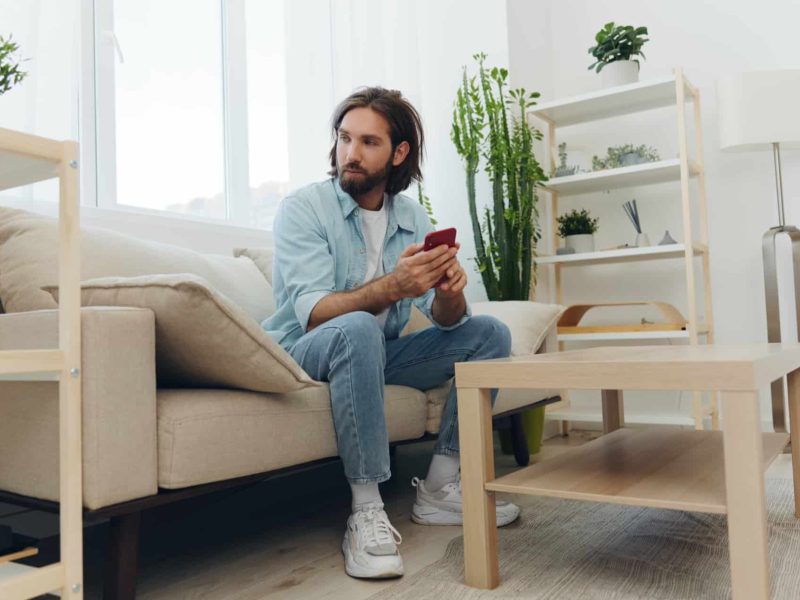 A man with a beard sits on the couch during the day and smiles at home and looks at his phone relaxing on his day off, life online on his phone texting with friends. High quality photo